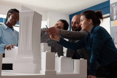 Group of multi ethnic colleagues work in architecture at project with blueprint plans building model maquette. Professional partners discussing vision of construction design on paper
